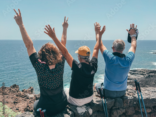 Back view of three friends in outdoor excursion at sea, sitting on the cliff looking at the horizon over water with open arms, enjoying freedom and vacation, healthy activity in retirement