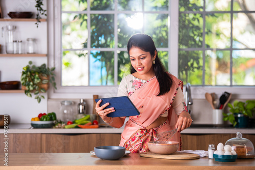 Indian young woman busy preparing recipe in kitchen from tablet pc