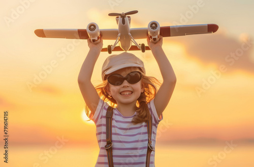 A happy child girl in striped Tshirt and pilot's cap holds up an airplane toy against the background of sunset sky, happy laughing kid playing with wooden plane at summer evening © Kien