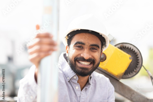 A smiling man in a white hard hat leans on a metal pole at a construction site. photo