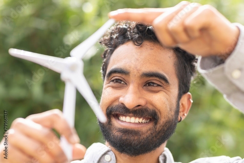 A smiling man with curly hair holds a small white wind turbine model above his head against a blurred green foliage background. photo
