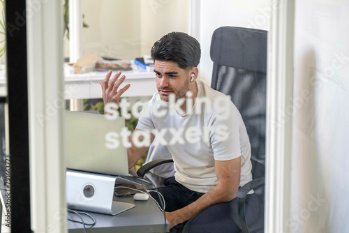 A man is sitting at a desk in a well-lit room, looking at a computer screen with a perplexed expression and gesturing with his hand. photo