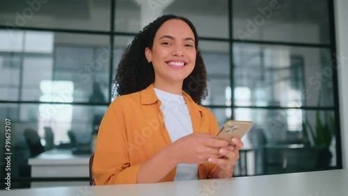 Amazing happy hispanic or brazilian curly haired woman in an orange shirt, sitting at a workplace in the office, using her mobile phone, messaging with friends, browsing social media, smile at camera photo