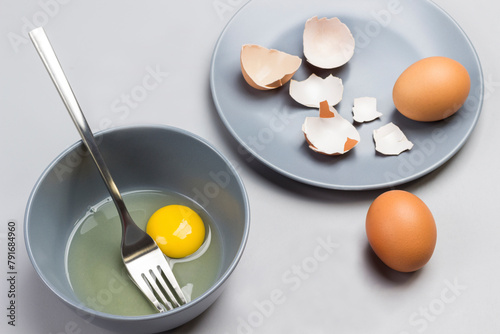 Broken egg and fork in gray bowl. Chicken shells on gray plate. Two brown eggs on table
