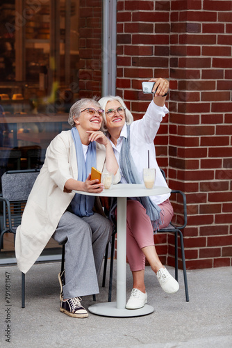 Sitting Outdoor Cafe stylish mature women with smiles capturing joyful selfie moment together, wearing glasses and smart casual attire, with drinks on table, relaxed social outing of elderly people