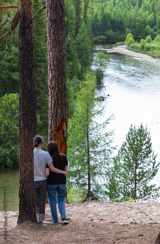 A couple of tourists admires a beautiful river and green forest from high steep bank on sunny day. Summer hikes and travels. River Ekhe-Ukhgun, foothill Tunka valley, Buryatia, Baikal region, Siberia photo