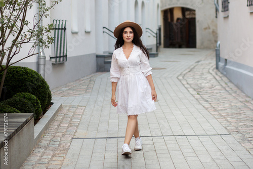 Girl in a hat and white dress on the street. Walking woman with long wavy hair in hat. Girl in a white dress and sneakers. Portrait of the young woman. Sensual portrait of a beautiful woman.