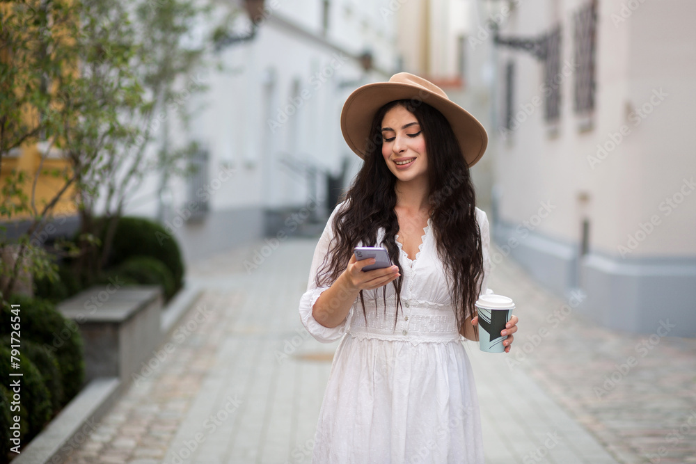 Girl in a hat and white dress on the street. Young  girl with coffee. Woman with long wavy hair in hat.  Girl in a white dress and sneakers. Girl in the city takes a selfie.