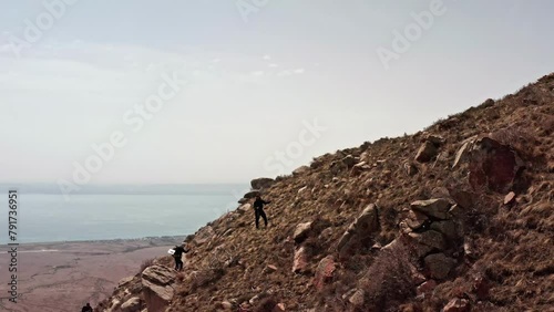 Climbers walking up a rocky slope to the top of a mountain, climbing over stones against the backdrop of a picturesque landscape and a lake in the distance. photo
