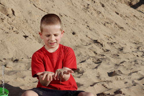 A six-year-old boy with short hair looks at the sand flowing through his fingers on a sunny summer day. Concept of summer holidays