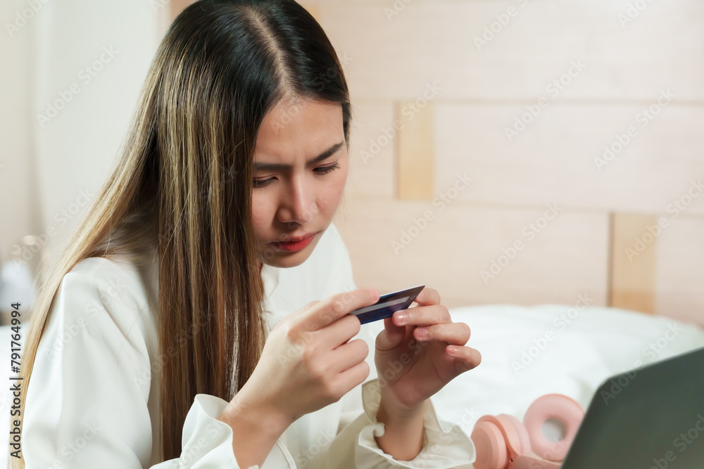 Asian lady in white, inspects credit card closely, troubled or questioning, in bedroom setting, laptop nearby, reflects concern over finances, security, or purchase. Concept work from home. Asian lady in white, inspects credit card closely, troubled or questioning, in bedroom setting, laptop nearby, reflects concern over finances, security, or purchase. Concept work from home.