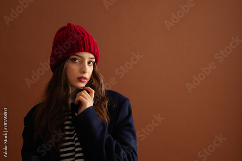 Fashionable confident woman wearing stylish red knitted beanie hat, sweater, classic blue woolen coat, posing on brown background. Studio fashion portrait. Copy, empty, blank space for text 
