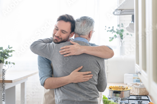 Cute loving caring adult caucasian son embracing hugging his old elderly senior father in the kitchen while cooking lunch, dinner, preparing meal together. Happy father`s day! I love you, dad! photo