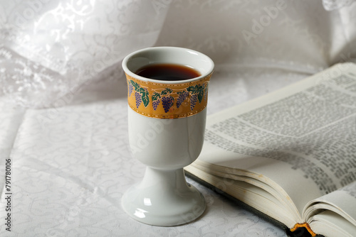 Porcelain goblet filled with wine resting on an open book. The goblet is adorned with a decorative band featuring grape illustrations. The scene is set against elegant, white lace curtains tablecloth