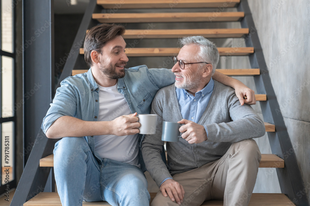 Adult caucasian young son supporting and hugging embracing his old senior elderly father drinking tea together sitting on the stairs at family home. Happy father`s day!