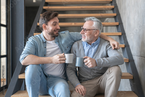 Adult caucasian young son supporting and hugging embracing his old senior elderly father drinking tea together sitting on the stairs at family home. Happy father`s day!