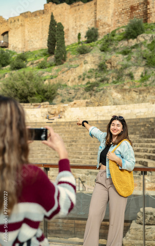 Two tourist friends in Malaga, one of them, with her back turned, focuses her camera on the other, who poses enthusiastically in front of a monument.