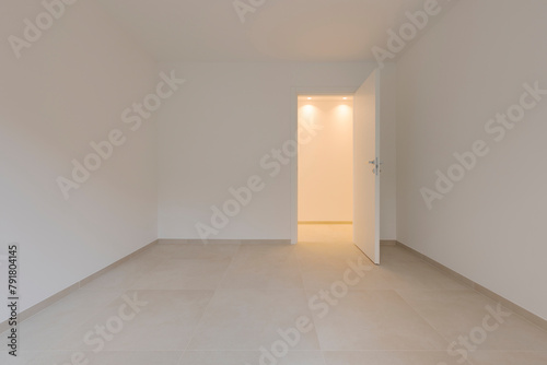 Inside an empty room and to the right a door leading to the corridor with a light on. All the walls and ceiling are white.