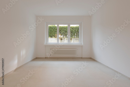 Empty interior of a room, with a central window in the background. Under the window a radiator.