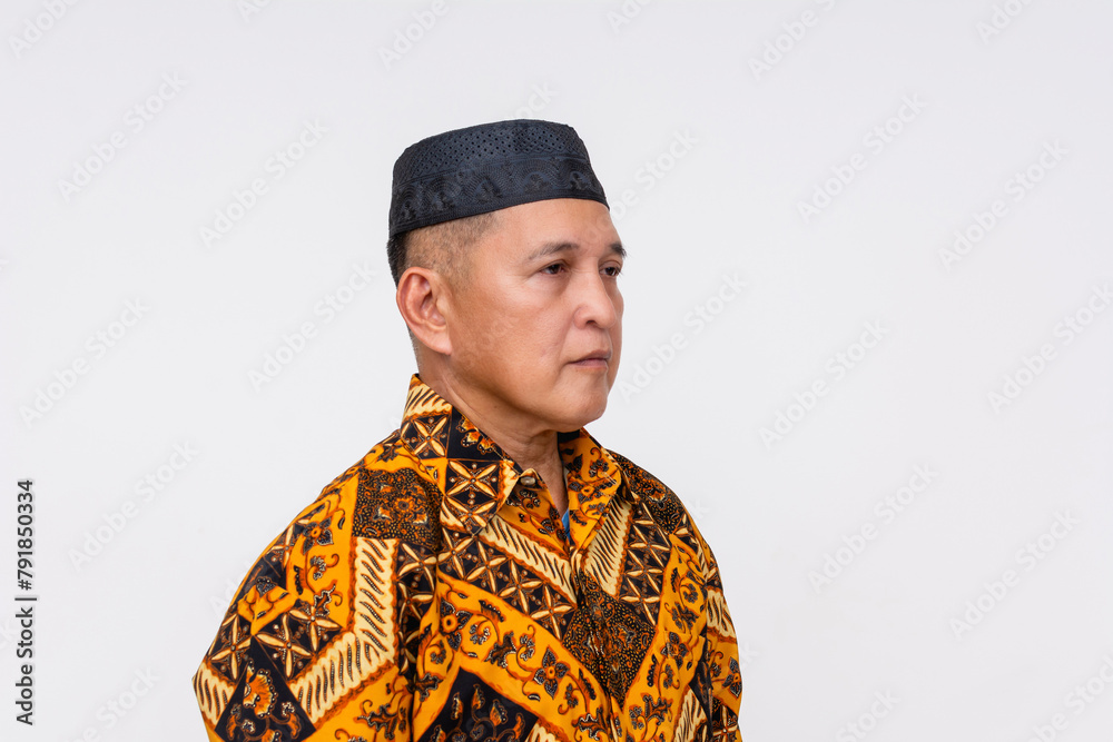 Portrait of a sober Indonesian man wearing a traditional batik shirt and a songkok, against a white background. Facing 45 degree angle.