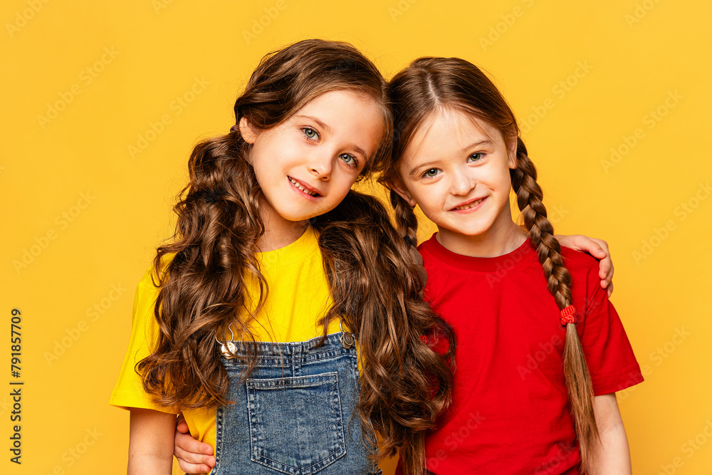 Two happy girls in bright summer clothes hugging and posing on a yellow background at studio. Summer children's fashion. Happy childhood. Advertising of children's products and sale Two happy girls in bright summer clothes hugging and posing on a yellow background at studio. Summer children's fashion. Happy childhood. Advertising of children's products and sale