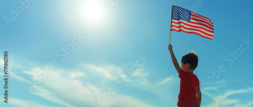 Back view of a young boy holding up the American flag against a bright blue sky, symbolizing hope, freedom, and patriotic spirit.