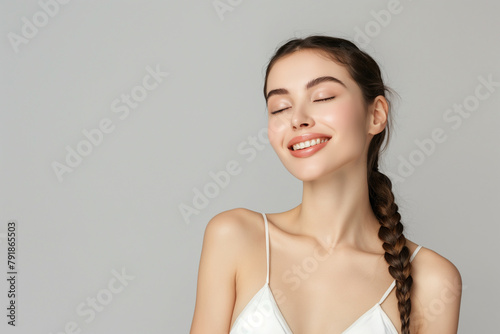 A serene young woman with a gentle smile and eyes closed, exuding peace and contentment with a sleek braid against a soft gray backdrop.