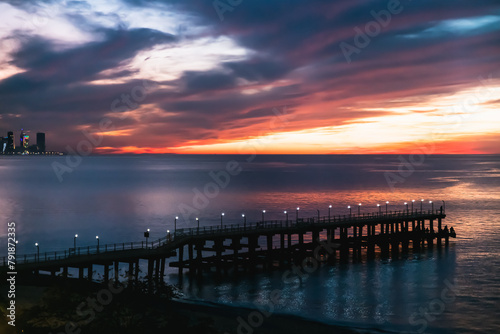 A pier illuminated by lanterns overlooking the sea, dramatic cumulus clouds in the red and blue colors of the sunset and downtown Batumi with high-rise buildings and night lights photo
