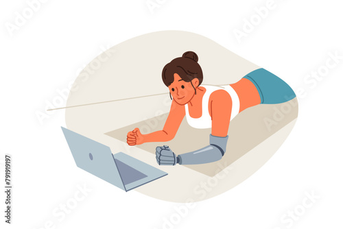 Sports woman with prosthetic arm trains at home and watches video lesson on laptop, lying on floor in plank. Sports girl doing yoga on fitness mat, training to improve health and muscle development