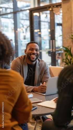A man is smiling and sitting at a table with a laptop