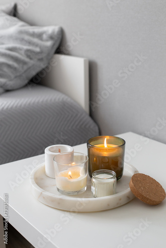 Home decor, stylish interior. Burning candles on marble tray standing on white badside table in bedroom. Bed with grey blanket and pillows.