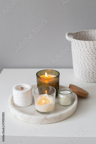 Home decor, stylish interior. Burning candles on marble tray and white wicker basket standing on white badside table in bedroom.