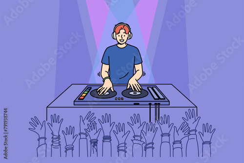 Man DJ performs in nightclub spinning records on mixing console, near crowd of partygoers with hands raised. Cheerful guy DJ in headphones plays dance hits for party or festival visitors