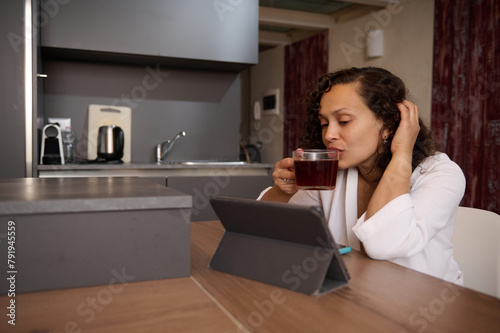 Beautiful woman in white bathrobe, sitting at kitchen table and drinking tea, watching movie or webinar on digital tablet