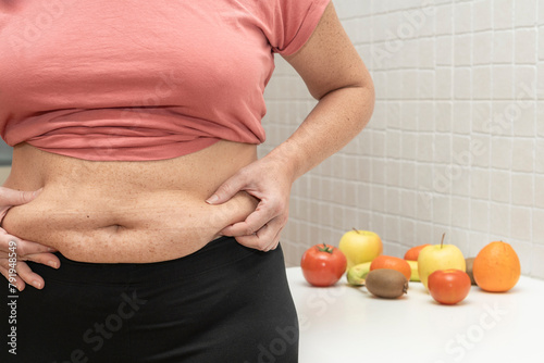 Obraz na plátně Middle-aged woman shows excess abdominal fat and in the background various fruits and vegetables