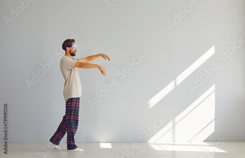 Full length portrait of young sleepwalker man in pajamas suffering from sleepwalking in sleep mask isolated on gray wall background. Somnambulism, insomnia and sleep problems concept. photo