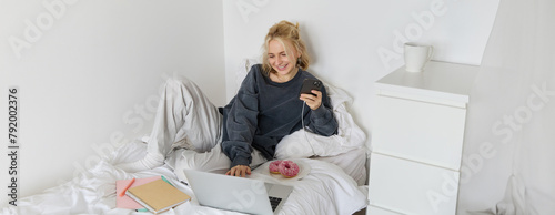 Lifestyle and people concept. Young beautiful woman, staying at home, lying in bed with laptop and smartphone, eating doughnut, enjoying free time, spending weekend at home, watching movie online