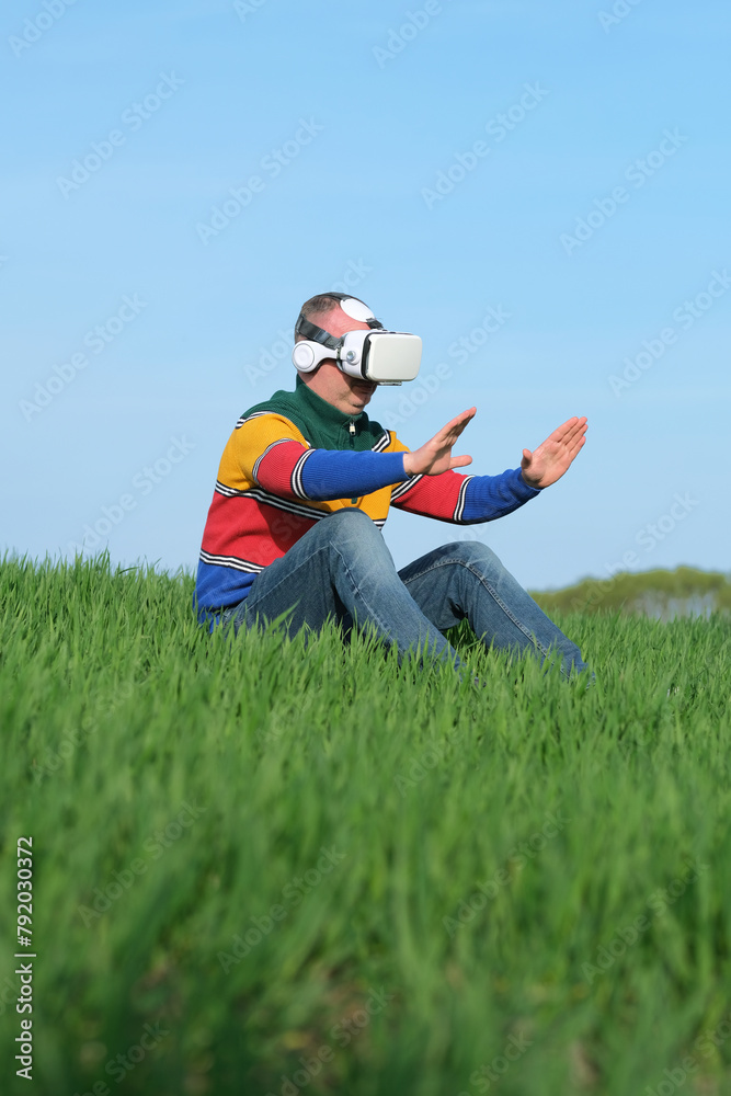 Amazed man touching air during the VR experience sitting on green grass in virtual reality glasses, side view. Sustainable agriculture. outdoors. Amazed man touching air during the VR experience sitting on green grass in virtual reality glasses, side view. Sustainable agriculture. outdoors.