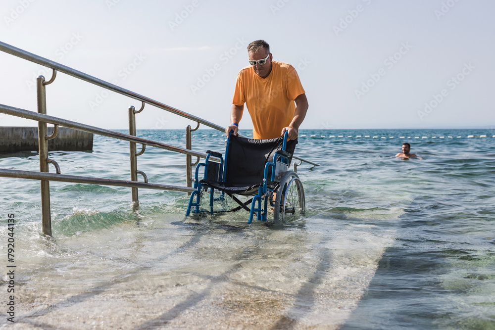 Male assistant helping a man in a wheelchair to enjoy the sea on an access ramp into the water. Accessible beach concept. Male assistant helping a man in a wheelchair to enjoy the sea on an access ramp into the water. Accessible beach concept.