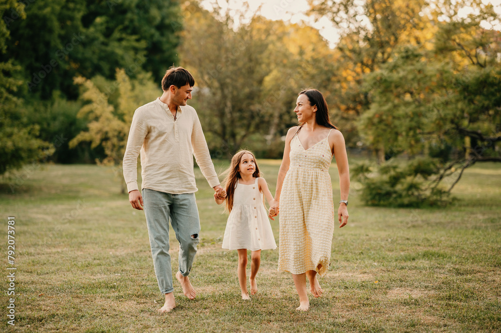 Cheerful young family of three walk together barefoot on grass while holding hands in park.