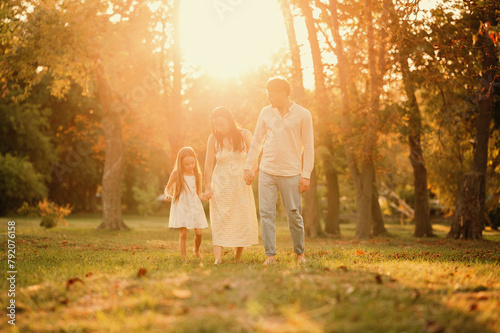 Wonderful young family of three are walking barefoot on grass while holding hands at sunset.