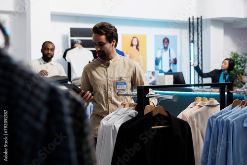 Clothing store manager using digital tablet and organizing merchandise to attract customers. Shopping center employee managing inventory and examining apparel hanging on rack