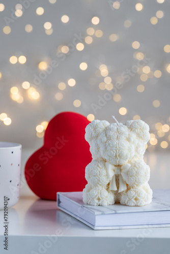 Handmade candle in the shape of teddy bear on white table with red heart and lights in background. Cute holiday gift. Soy wax candles. Valentine's day present