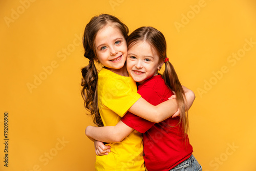 Two happy girls in bright summer clothes hugging and posing on a yellow background at studio. Summer children's fashion. Happy childhood. Advertising of children's products and sale