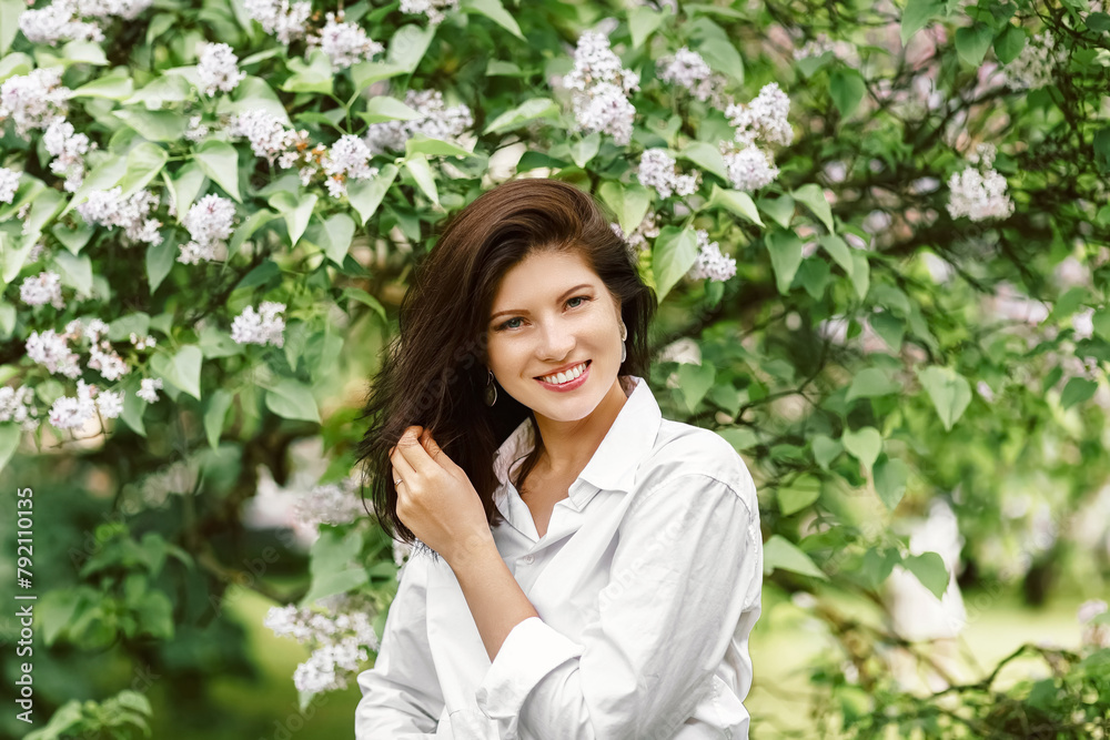 Face and cute smile wearing casual white blouse or shirt in the garden among blooming apple tree branches with small flowers and green leaves looking at camera in sunny day Face and cute smile wearing casual white blouse or shirt in the garden among blooming apple tree branches with small flowers and green leaves looking at camera in sunny day