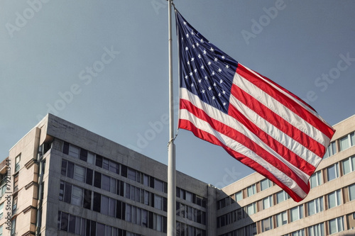 The image shows a large American flag waving in front of a government building. The flag is blowing in the wind and the sun is shining on it. The building is made of concrete and has a lot of windows