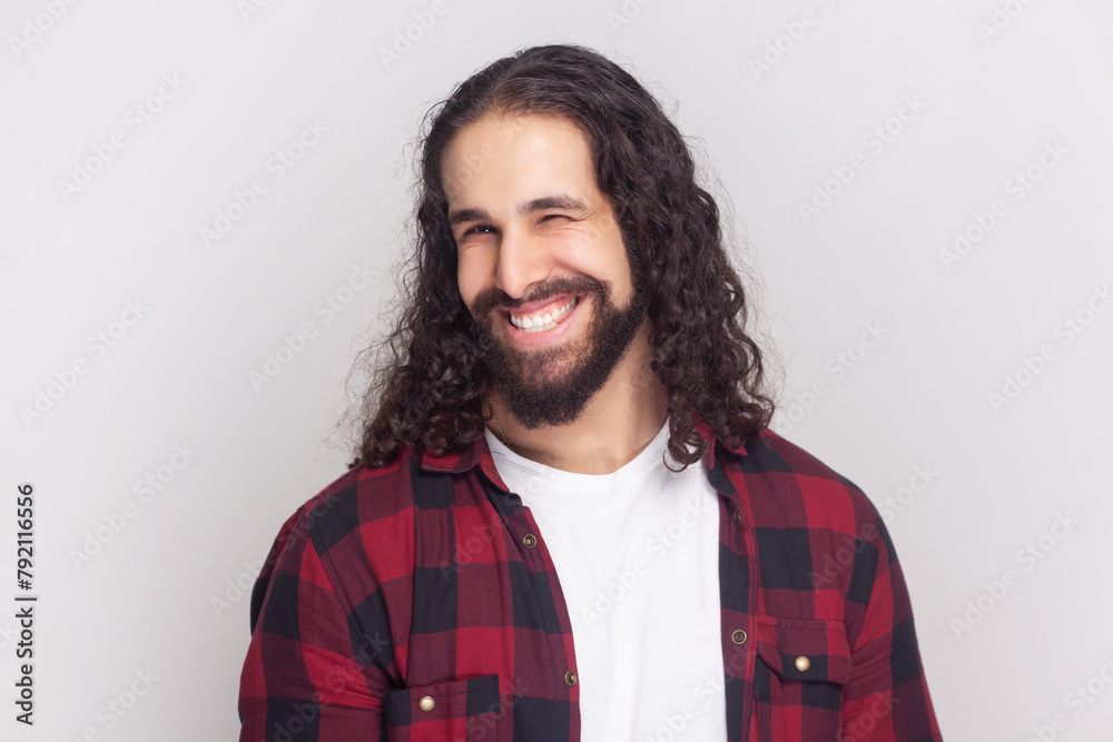 Bearded man with long curly hair in checkered red shirt winking while having good mood smiling, blinking eye showing his sympathy or positiveness. Indoor studio shot isolated on gray background. Bearded man with long curly hair in checkered red shirt winking while having good mood smiling, blinking eye showing his sympathy or positiveness. Indoor studio shot isolated on gray background.