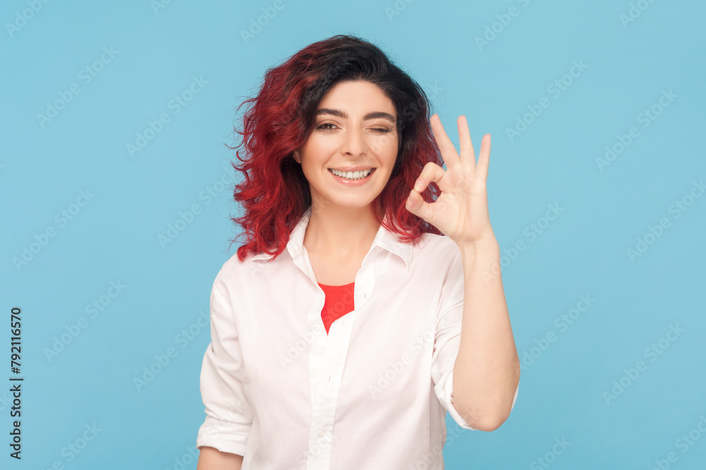 Portrait of cheerful adult woman with fancy red hair showing okay sign, assures you everything is fine, looks gladly, wearing white shirt. Indoor studio shot isolated on blue background. Portrait of cheerful adult woman with fancy red hair showing okay sign, assures you everything is fine, looks gladly, wearing white shirt. Indoor studio shot isolated on blue background.