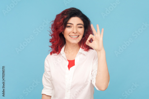 Portrait of cheerful adult woman with fancy red hair showing okay sign, assures you everything is fine, looks gladly, wearing white shirt. Indoor studio shot isolated on blue background. photo