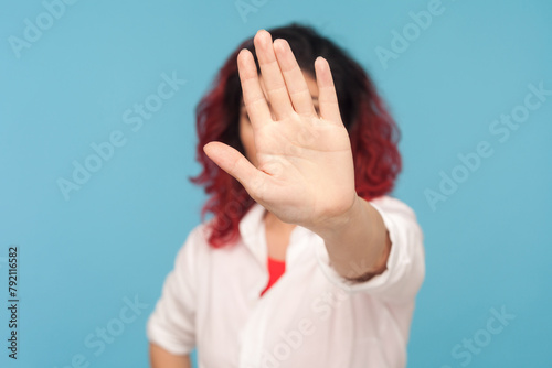 Portrait of unknown woman with fancy red hair showing stop sign, demonstrating prohibition gesture, feels danger, wearing white shirt. Indoor studio shot isolated on blue background.